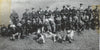 A black-and-white photo from "The Fighting Fathers" (Schiffer Publishing, Ltd.) shows soldiers—some linked to CIA South Vietnam ops—posing outdoors with rifles or submachine guns in mixed uniforms and hats, standing, kneeling, or prone on the grass.
