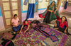 Four women in traditional attire, representing Indian artisans, craft and converse among vibrant embroidered textiles. Artisans by Design by Schiffer Publishing, Ltd. captures their creativity in a room full of colorful handmade beauty.