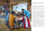 A group of adults and children gather around a low table at Somaiya Kala Vidya as a woman in blue assists, celebrating traditional artisans of India, featured in "Artisans by Design" from Schiffer Publishing, Ltd. A whiteboard is seen behind them.