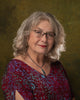 An older woman with wavy gray hair, glasses, a red patterned top, and a silver necklace with blue pendant poses like a Boadicea's Tarot of Earthly Delights card by Schiffer Publishing, Ltd. against a mottled green and brown backdrop.