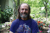 An older man with long hair, a beard, and glasses in a colorful horse t-shirt smiles in a lush garden, perhaps reflecting on quotes from The Law of One Contemplation Deck by Schiffer Publishing, Ltd. among blooming flowers on a sunny day.
