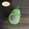 The Crochet Your Veggies Amigurumi Kit Avocado by Schiffer Publishing, Ltd. features a smiling crocheted avocado with a textured pit sitting on wood beside scattered soil. The kit includes everything you need to make 1 avocado.