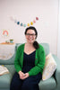 A woman with dark curly hair and glasses smiles on a light blue couch, with pastel felt wall decor behind her. Featured: "HeartFELT Crafts to Make and Give" by Schiffer Publishing, Ltd., inspiration for handmade home accents.