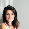 A woman with long dark hair and light skin, in a white top, sits by a sunlit gray background, gently smiling at the camera. She embodies the essence of Ukrainian Spiritual Traditions by Schiffer Publishing, Ltd.