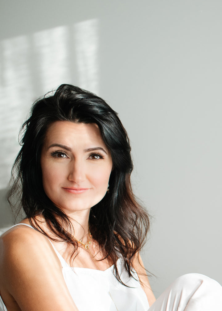 A woman with long dark hair and light skin, in a white top, sits by a sunlit gray background, gently smiling at the camera. She embodies the essence of Ukrainian Spiritual Traditions by Schiffer Publishing, Ltd.