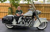 A man with glasses and a beard sits on a Gilroy Indians motorcycle by Schiffer Publishing, Ltd., parked in a driveway with a wooden fence, plants, and trees in the background.