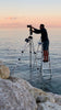 A man on a step ladder in shallow ocean water adjusts a camera at sunset, with rocks nearby and the calm sea under a pink sky—capturing scenes for Midwest Home by Schiffer Publishing, Ltd.