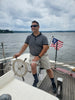 A man in sunglasses and casual attire steers a boat with an American flag on a cloudy day, capturing the spirit found in Schiffer Publishing, Ltd.'s "American Merchant Seaman's Manual, 8th Edition.