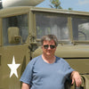 A man in sunglasses and a blue t-shirt stands outside on a sunny day beside an olive green Sd.Kfz. 251 half-track (Schiffer Publishing, Ltd.) with a white star. A portion of a missile is visible atop the vehicle.