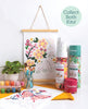 A ColorMaps Paint-by-Number Relaxing Garden display by Schiffer Publishing, Ltd. shows floral artwork, paint pots, brushes in a jar, a potted plant, rolled kits, and a completed pink flowers painting. A sign reads: Collect Both Kits!.