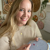 A smiling woman with long blonde hair holds the Sunny Stitches Embroidery Kit Sunny Days Ahead by Schiffer Publishing, Ltd., surrounded by embroidery art and supplies in a cozy, well-lit craft room.