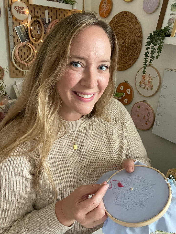 A woman with long blonde hair in a beige sweater smiles while working with a needle on the Sunny Stitches Embroidery Kit Woodland Mushroom by Schiffer Publishing, Ltd. in a cozy craft room decorated with embroidery hoops.