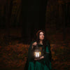 A woman in a green dress and flower crown sits on the forest floor at dusk, holding a glowing lantern amid autumn leaves and tall dark trees, evoking the rich symbolism of Schiffer Publishing, Ltd.’s The Tree Tarot.
