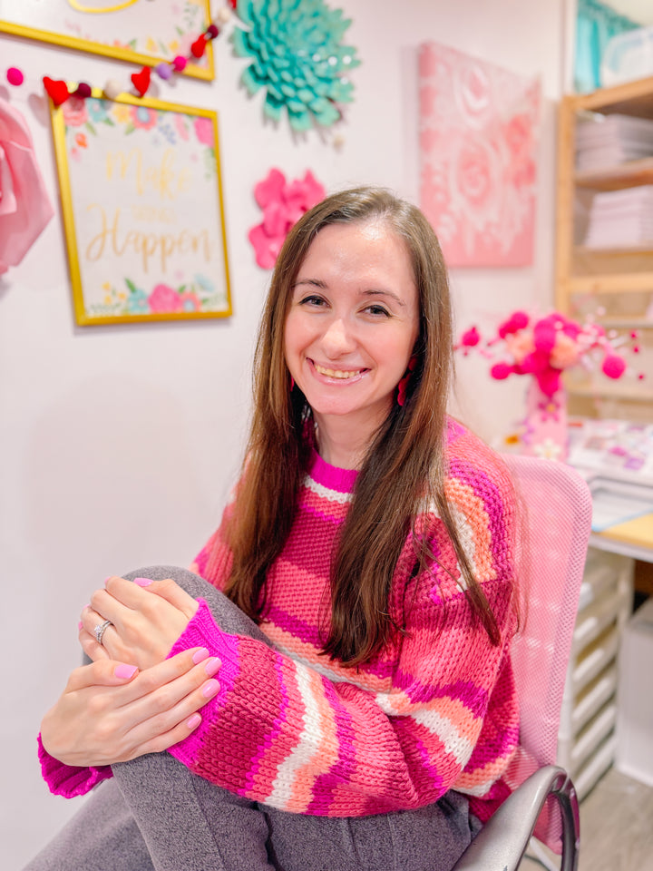 A woman with long brown hair, wearing a pink and red striped sweater, sits smiling in a colorful, craft-themed room adorned with art, paper flowers, cheerful signs, and whimsical stickers from Schiffer Publishing’s A Day in Cozy Town: The Bookshop Coloring and Sticker Book.
